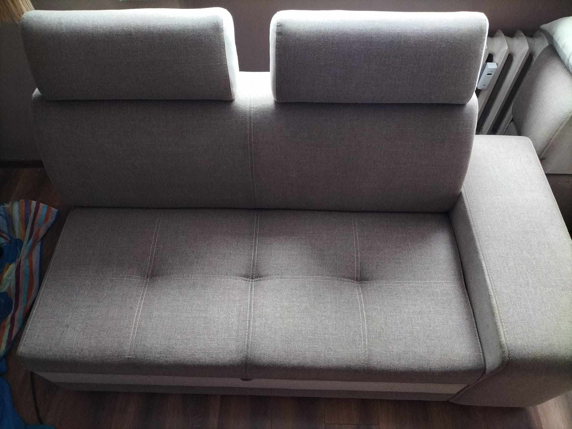 Szara, rozłożona kanapa z regulowanymi zagłówkami, widoczna z tyłu, z drewnianą podłogą i fragmentem grzejnika w tle.