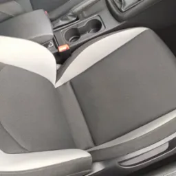 Szare i białe obicie fotela samochodowego z widocznym podłokietnikiem, konsolą środkową i gumową wycieraczką podłogową z geometrycznym wzorem.