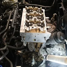 Widok z g&oacute;ry na silnik samochodowy w trakcie naprawy, z widoczną głowicą cylindr&oacute;w, wałkami rozrządu i elementami osprzętu, oświetlony lampą warsztatową.