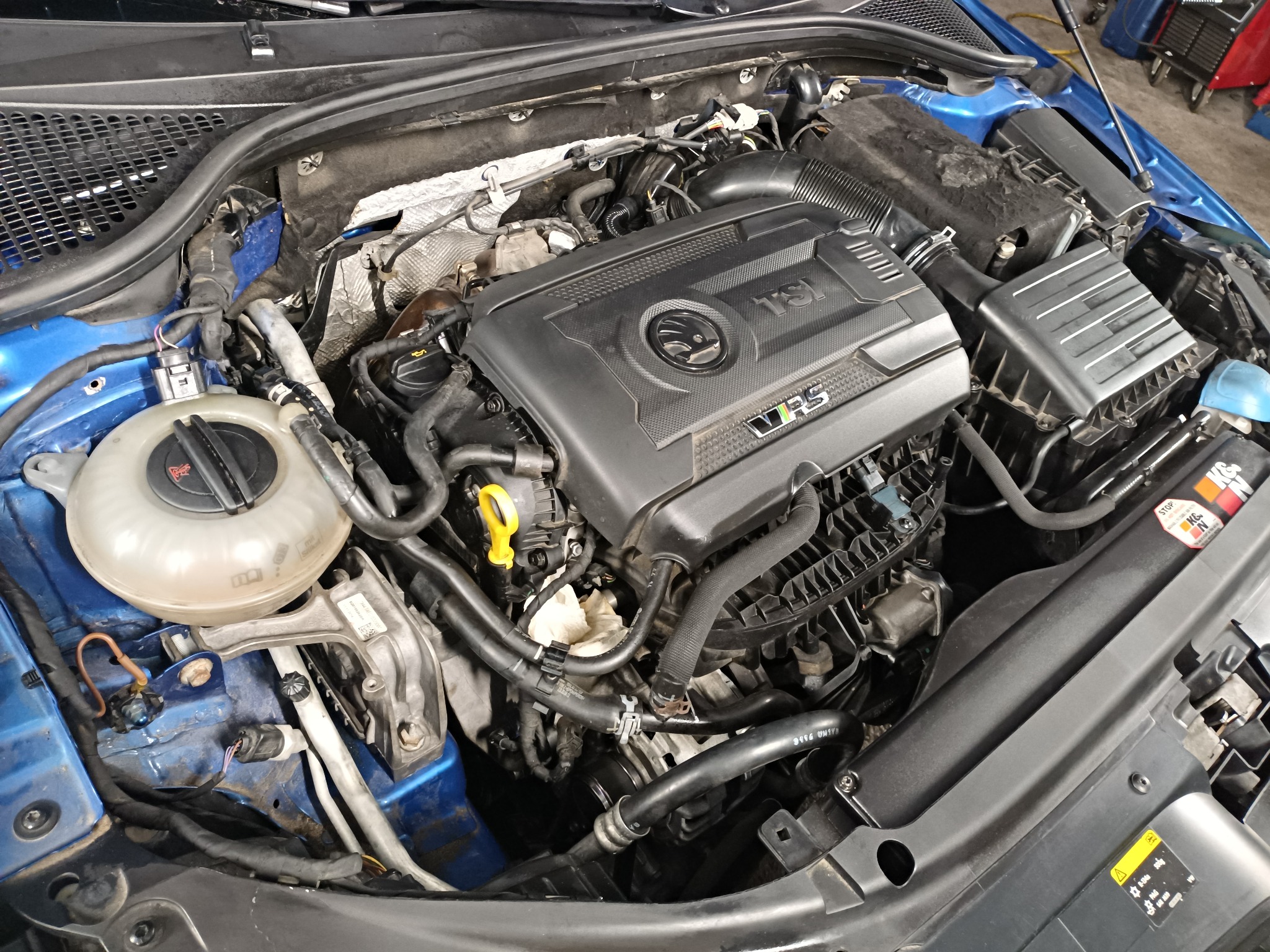 Otwarta maska błękitnego samochodu, widoczny silnik TSI z elementami osprzętu, w tym zbiornik wyrównawczy płynu chłodniczego i liczne przewody. Widoczny fragment karoserii w kolorze niebieskim.