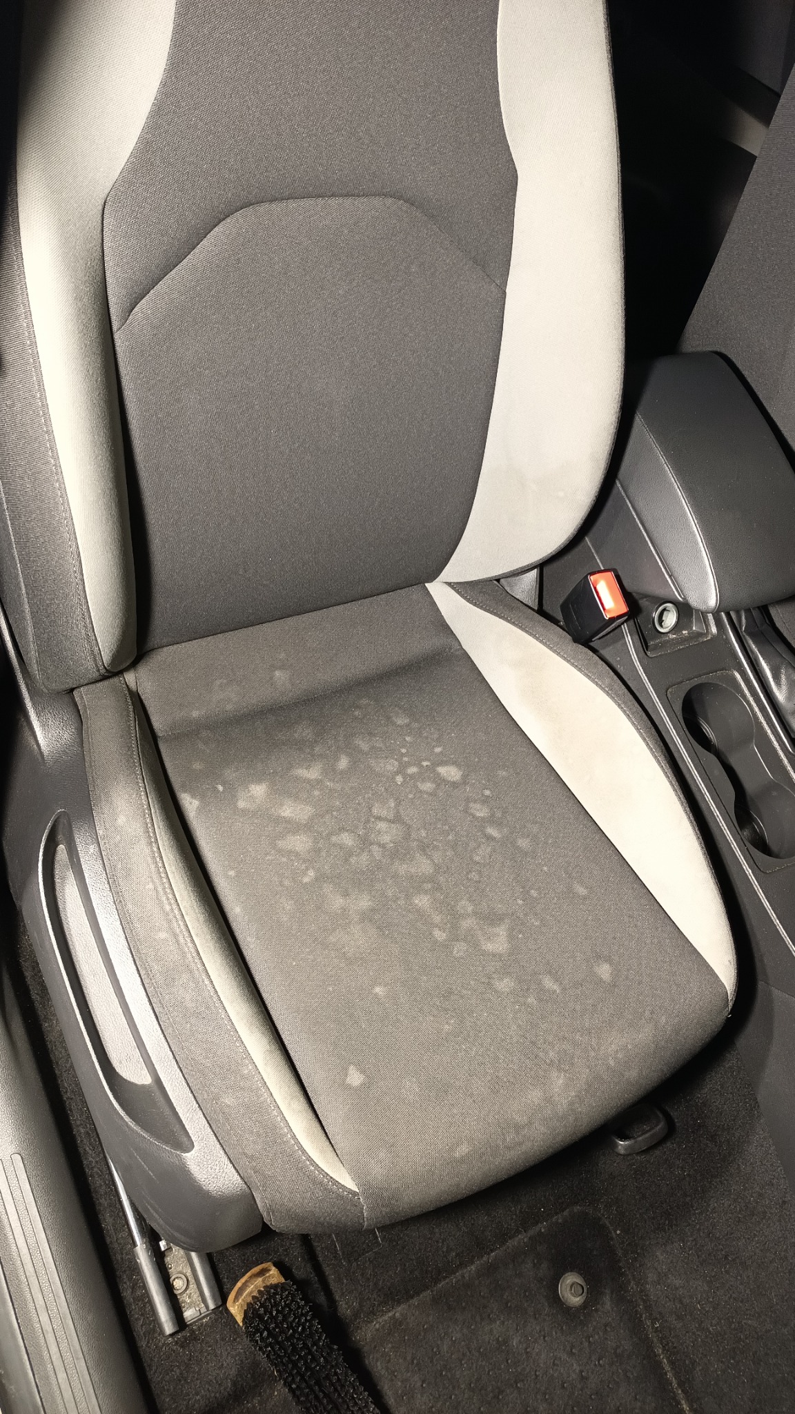 Szare samochodowe siedzenie z jasnymi wstawkami, widoczne plamy przed czyszczeniem, obok leży szczotka.