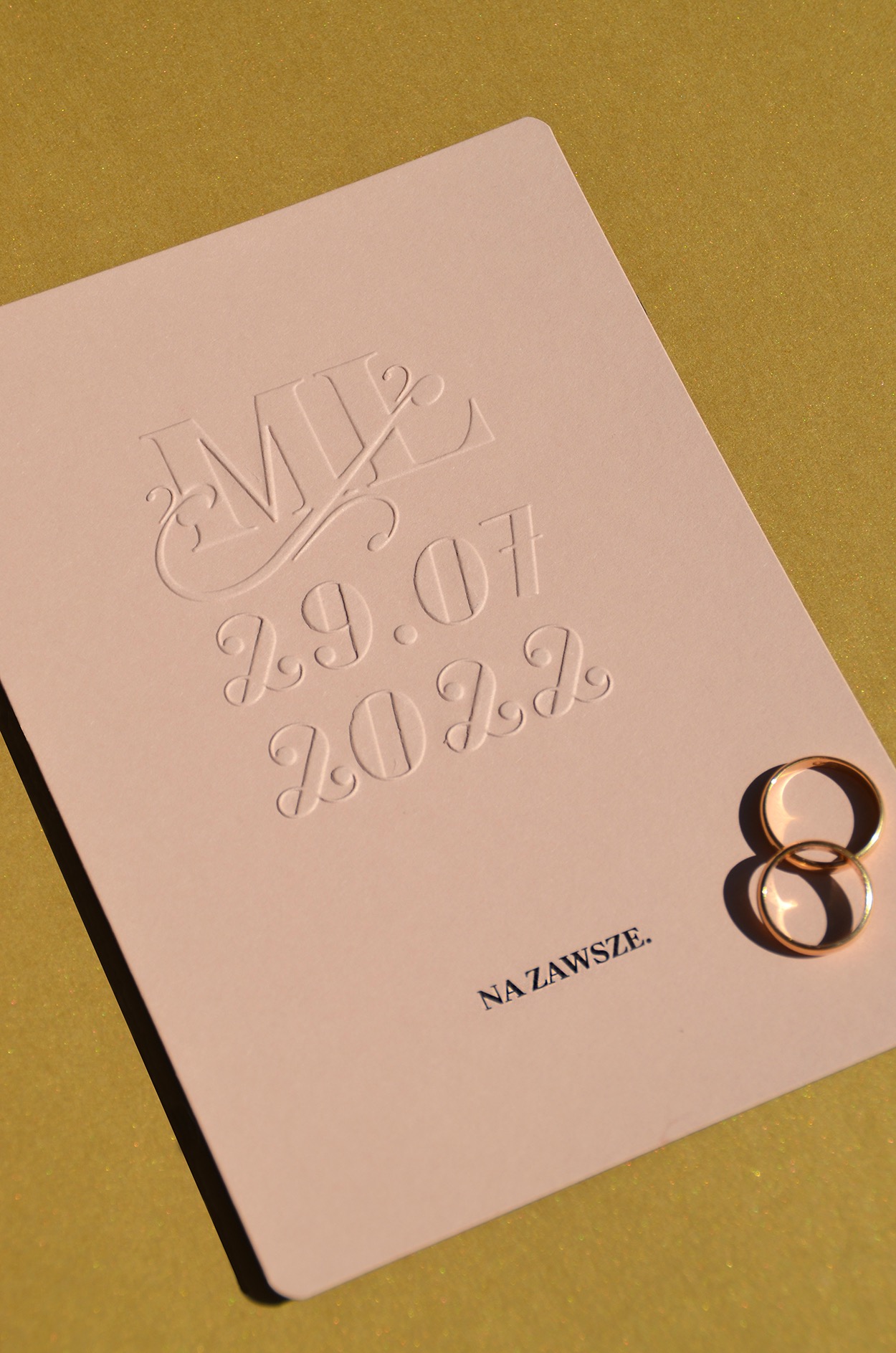 Bladoróżowa kartka z wytłoczonymi inicjałami, datą ślubu (29.07.2022) i napisem 'Na Zawsze', obok leżą dwie złote obrączki na złotym tle.