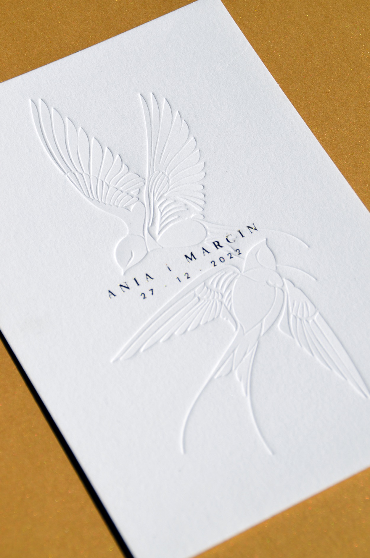 Elegancka, biała kartka z tłoczonym motywem dwóch jaskółek i napisem 'Ania i Marcin 27.12.2022' na złotym tle.