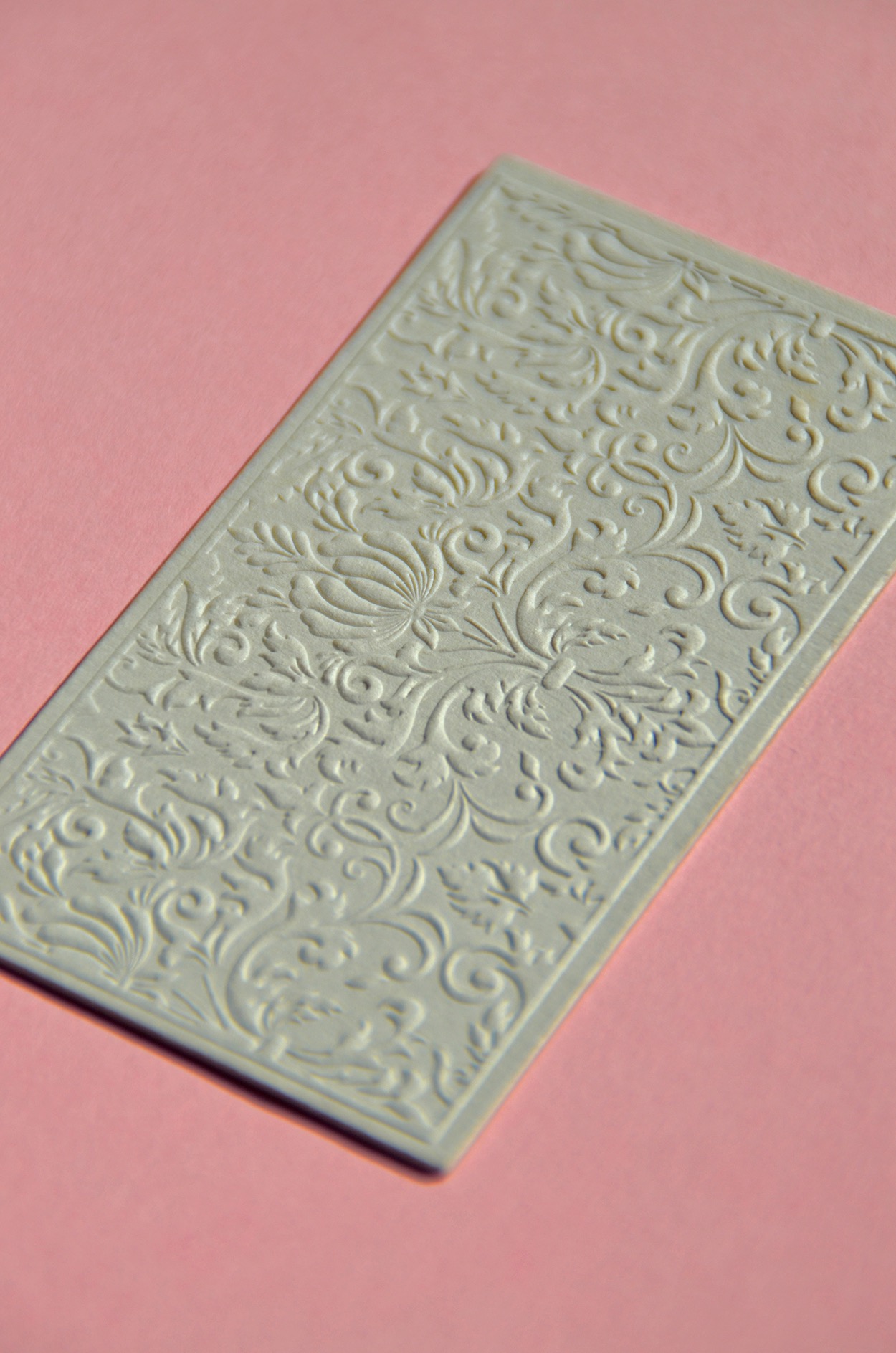 Elegancka wizytówka z wytłaczanym, roślinnym wzorem na różowym tle.