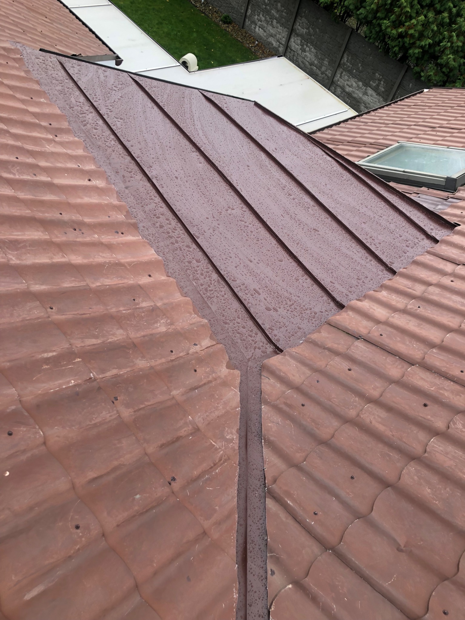 Brązowy dach z dachówki ceramicznej i blachy trapezowej, pokryty kroplami deszczu, widok z góry na fragment dachu z oknem dachowym.