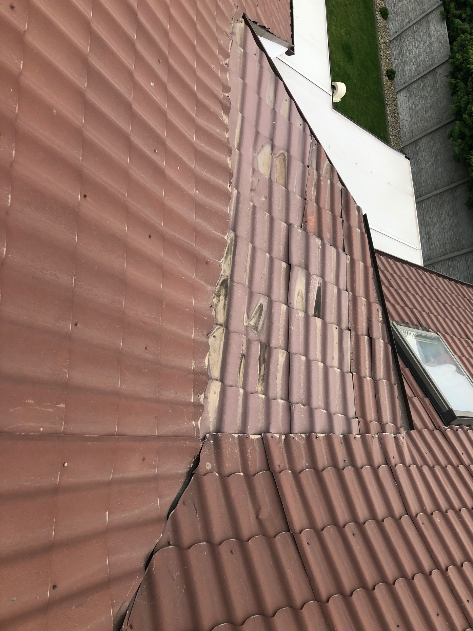 Uszkodzony dach z brązowej dachówki ceramicznej, widoczne ubytki i pęknięcia, fragment dachu z oknem dachowym, w tle budynek z białą elewacją i zieleń.