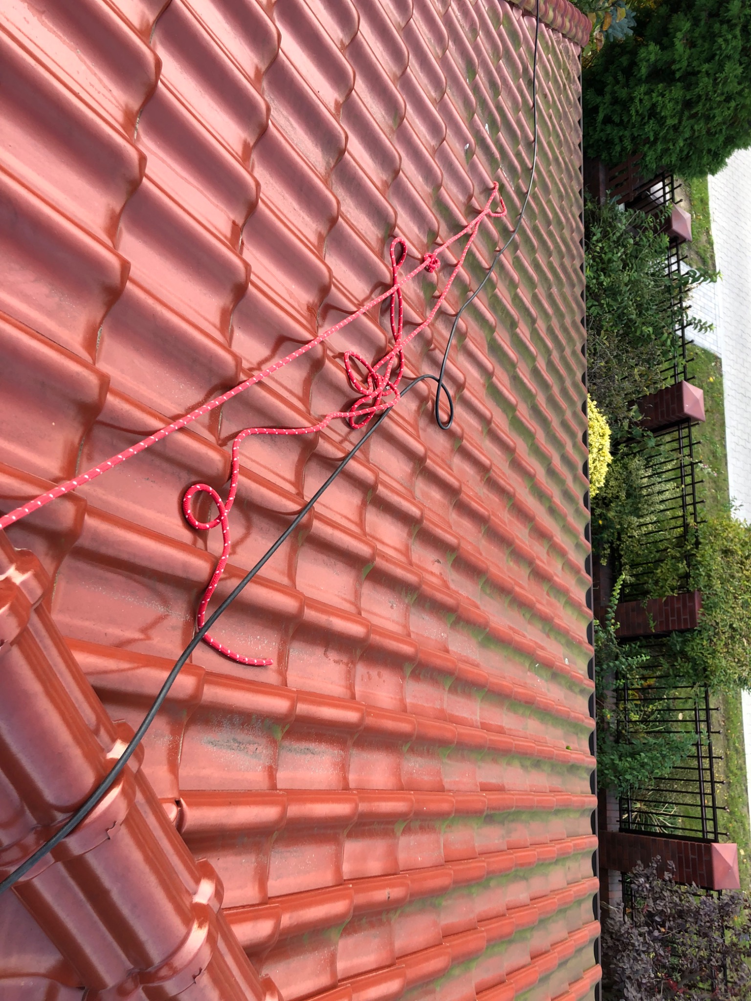 Czerwona dachówka ceramiczna z widocznymi śladami mchu i zamocowaną czerwoną liną z białymi kropkami, ujęcie z góry, widoczne elementy ogrodu w tle.