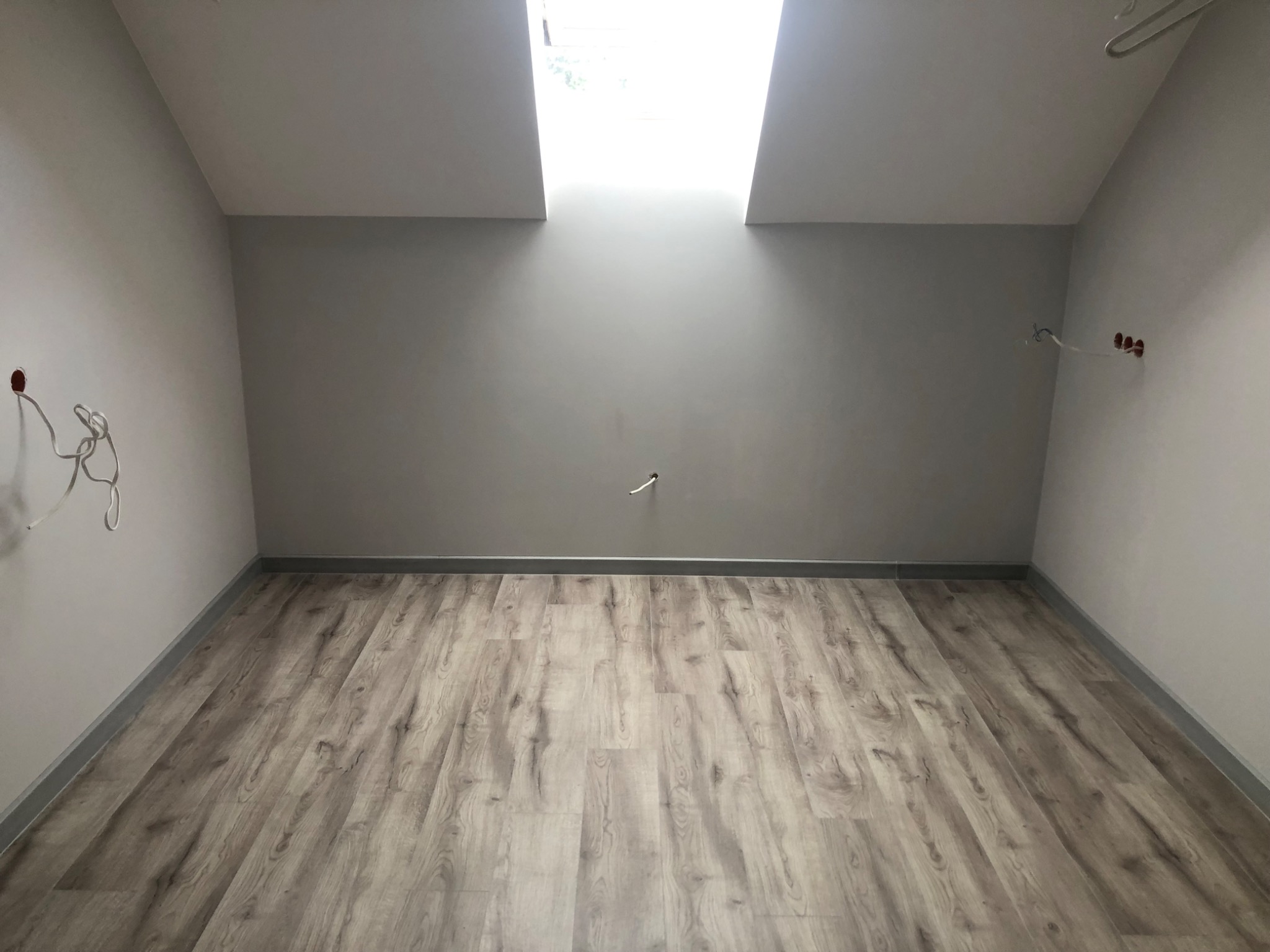Poddasze z oknami dachowymi, świeżo położone panele podłogowe z szarą listwą przypodłogową, ściany pomalowane na szaro, widoczne wystające kable elektryczne.
