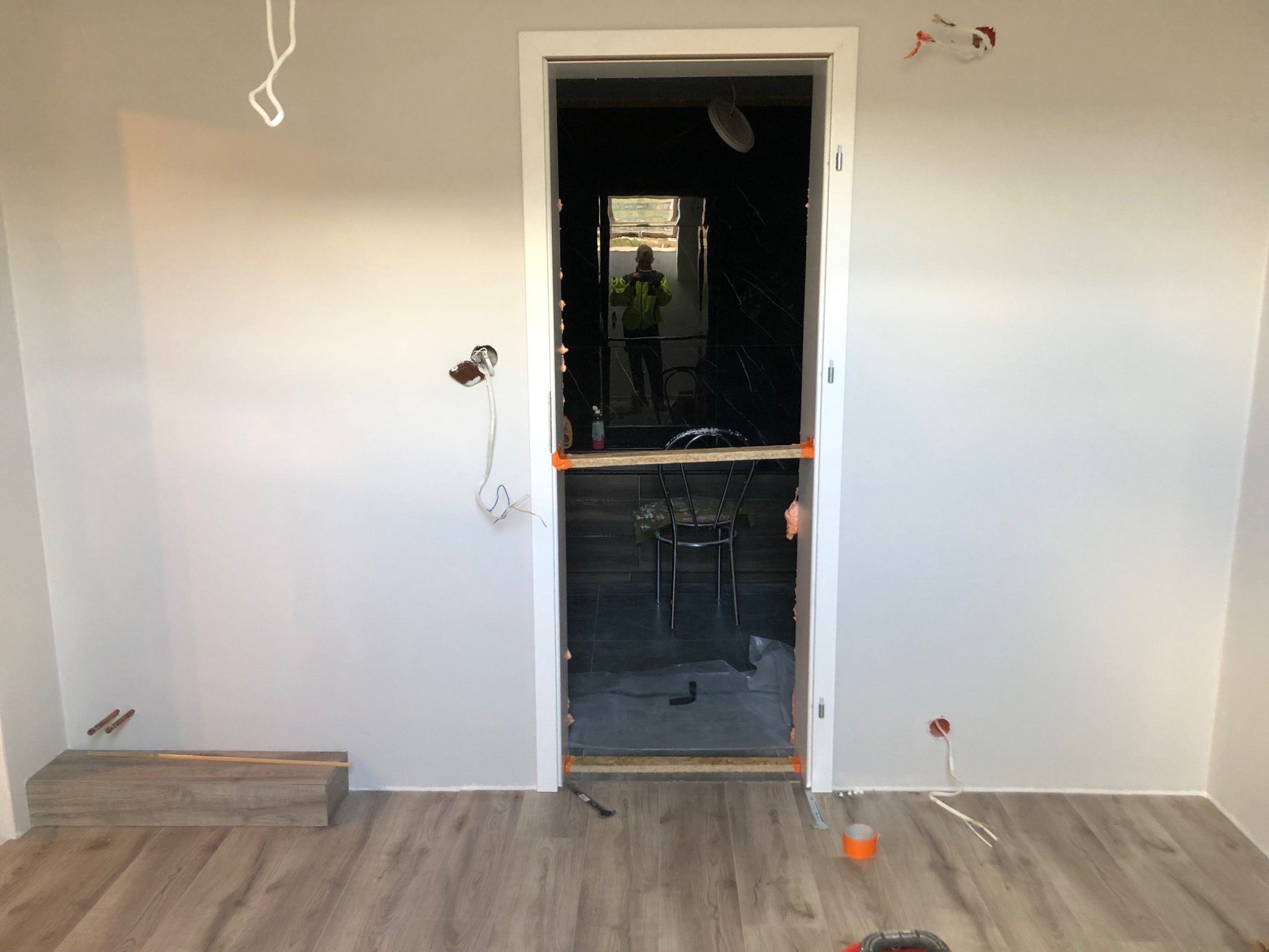 Wnętrze w trakcie remontu: widoczne przygotowania do położenia podłogi, fragment ułożonych paneli, ściany przygotowane do malowania, otwarty prześwit do pomieszczenia z czarnymi płytkami i odbiciem...