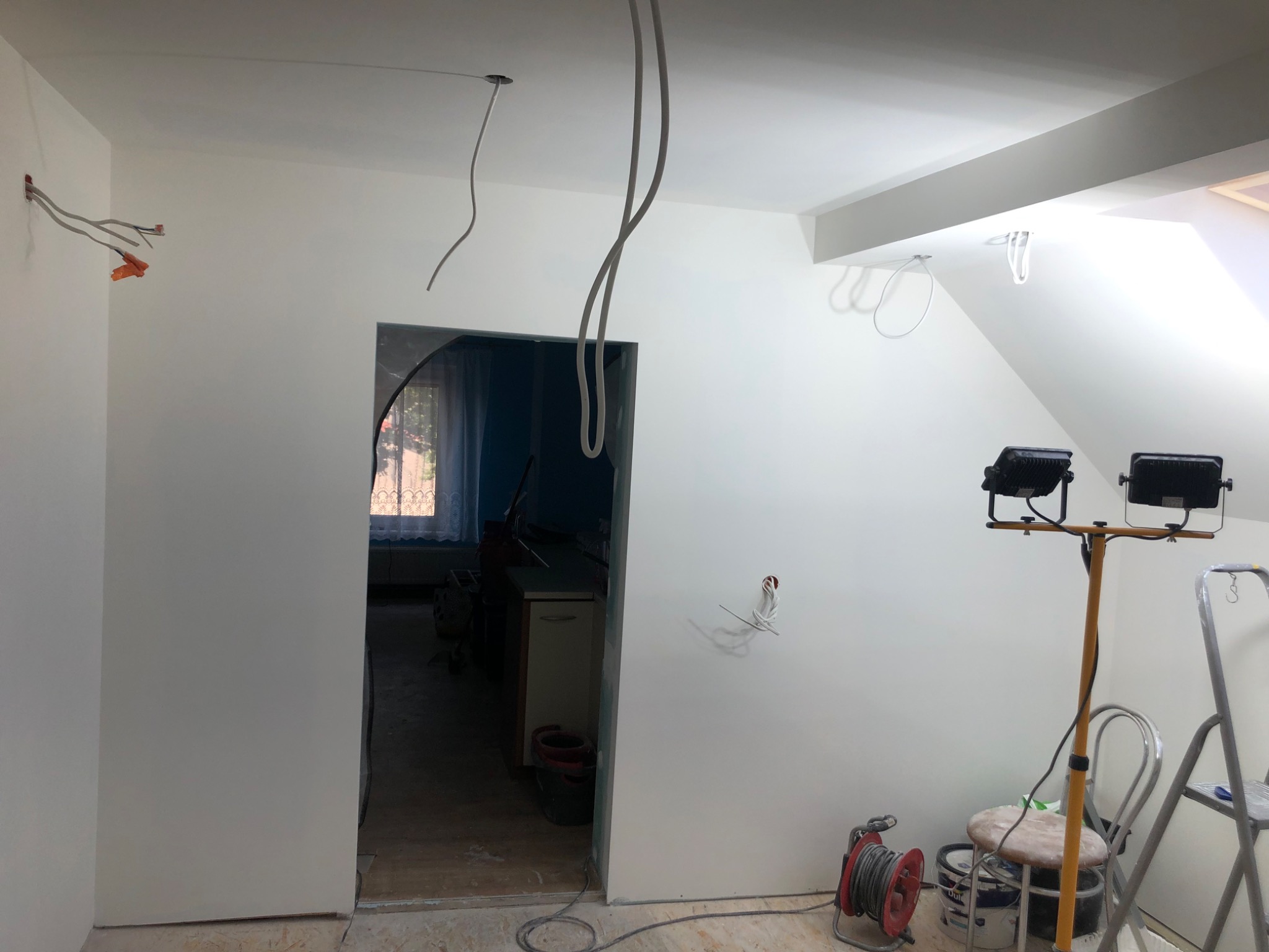Pokój w trakcie prac wykończeniowych: świeżo pomalowane ściany na biało, widoczne wystające przewody elektryczne, lampy budowlane na statywie, drabina, wiadro z farbą i przedłużacz.