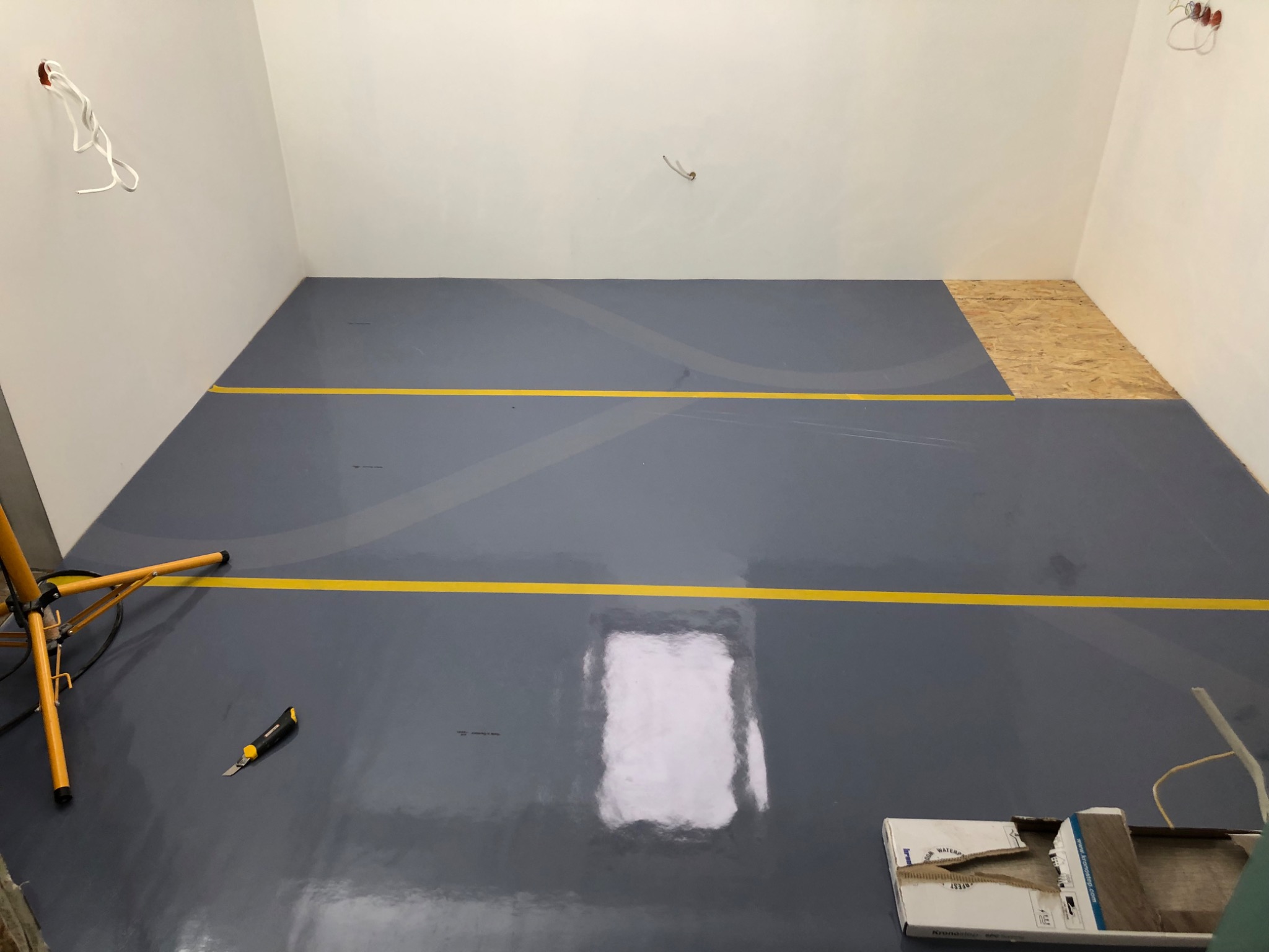 Przygotowanie podłoża pod panele: wyrównana, szara wylewka z żółtymi liniami wyznaczającymi obszar, fragment płyty OSB, lampa budowlana i nożyk w pomieszczeniu z białymi ścianami.