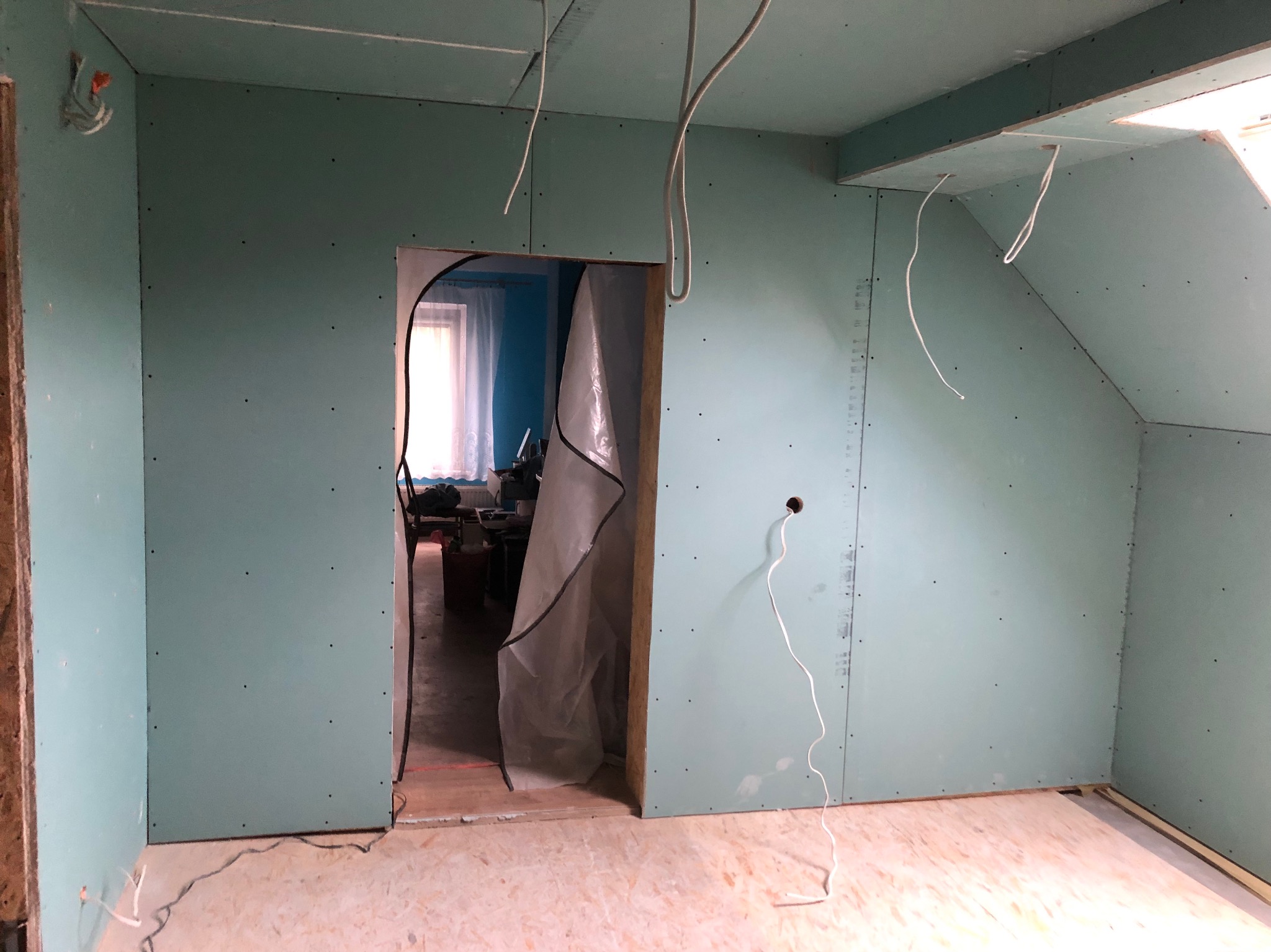 Wnętrze poddasza z zamontowanymi płytami gipsowo-kartonowymi, widoczne wystające przewody elektryczne i otwór drzwiowy zabezpieczony folią malarską.