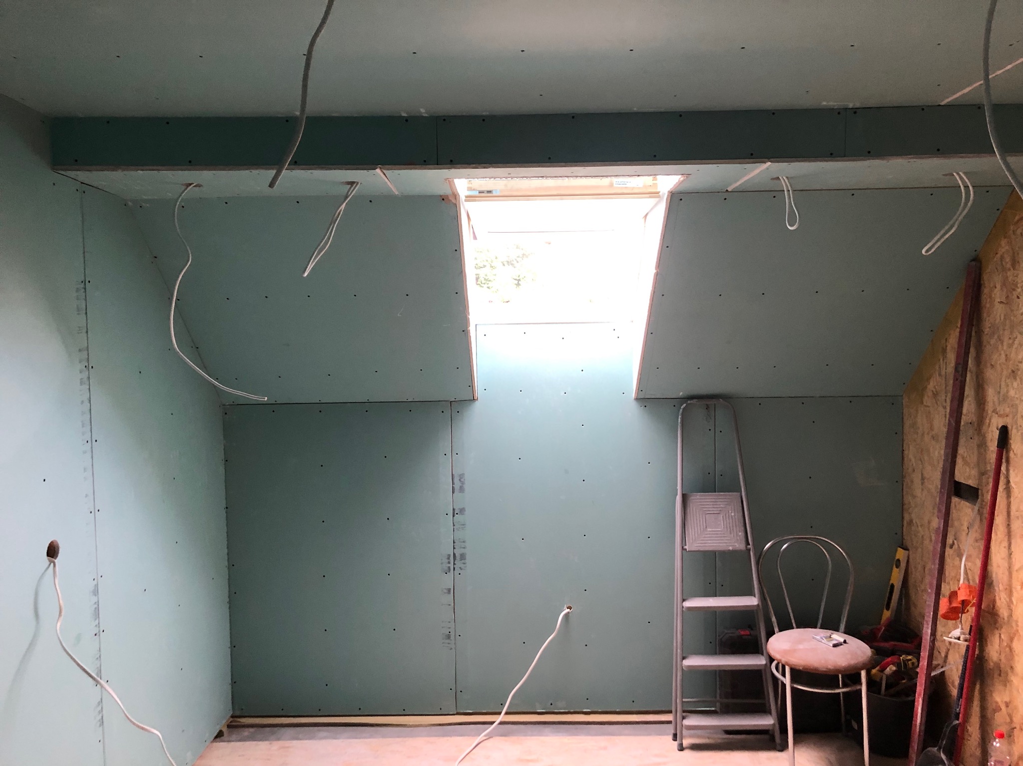 Wnętrze poddasza z zamontowanymi płytami kartonowo-gipsowymi, widoczne wystające kable, drabina aluminiowa, krzesło i narzędzia budowlane przy ścianie z desek.