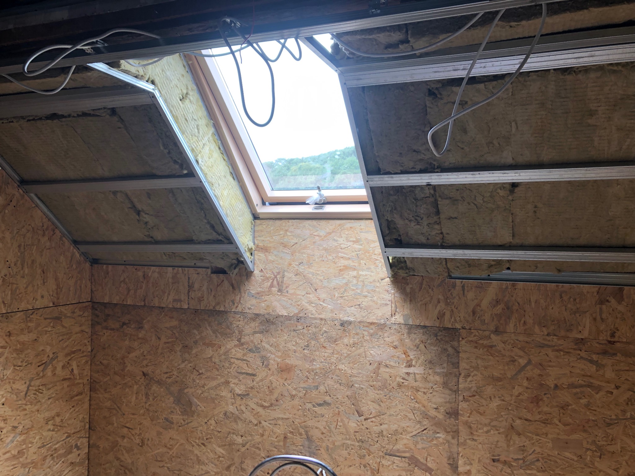 Montaż okna dachowego: widoczna izolacja wełną mineralną, metalowe profile konstrukcyjne, płyta OSB na ścianach poddasza, okno dachowe z widokiem na zieleń.