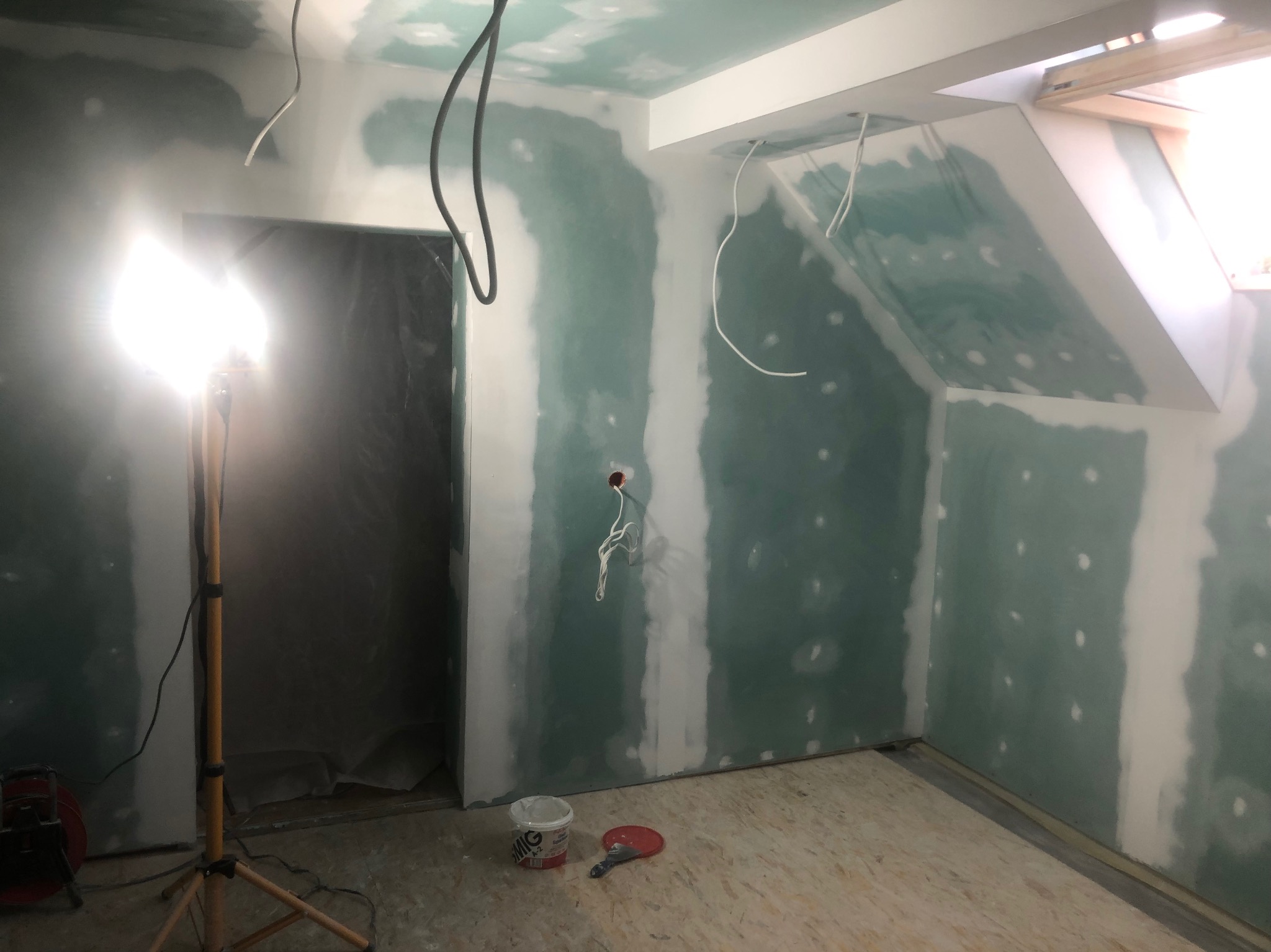 Pokój na poddaszu w trakcie remontu: ściany i sufit pokryte płytami kartonowo-gipsowymi, widoczne łączenia szpachlowane, oświetlenie halogenowe na statywie, kable zwisające z sufitu, wiadro z masą...