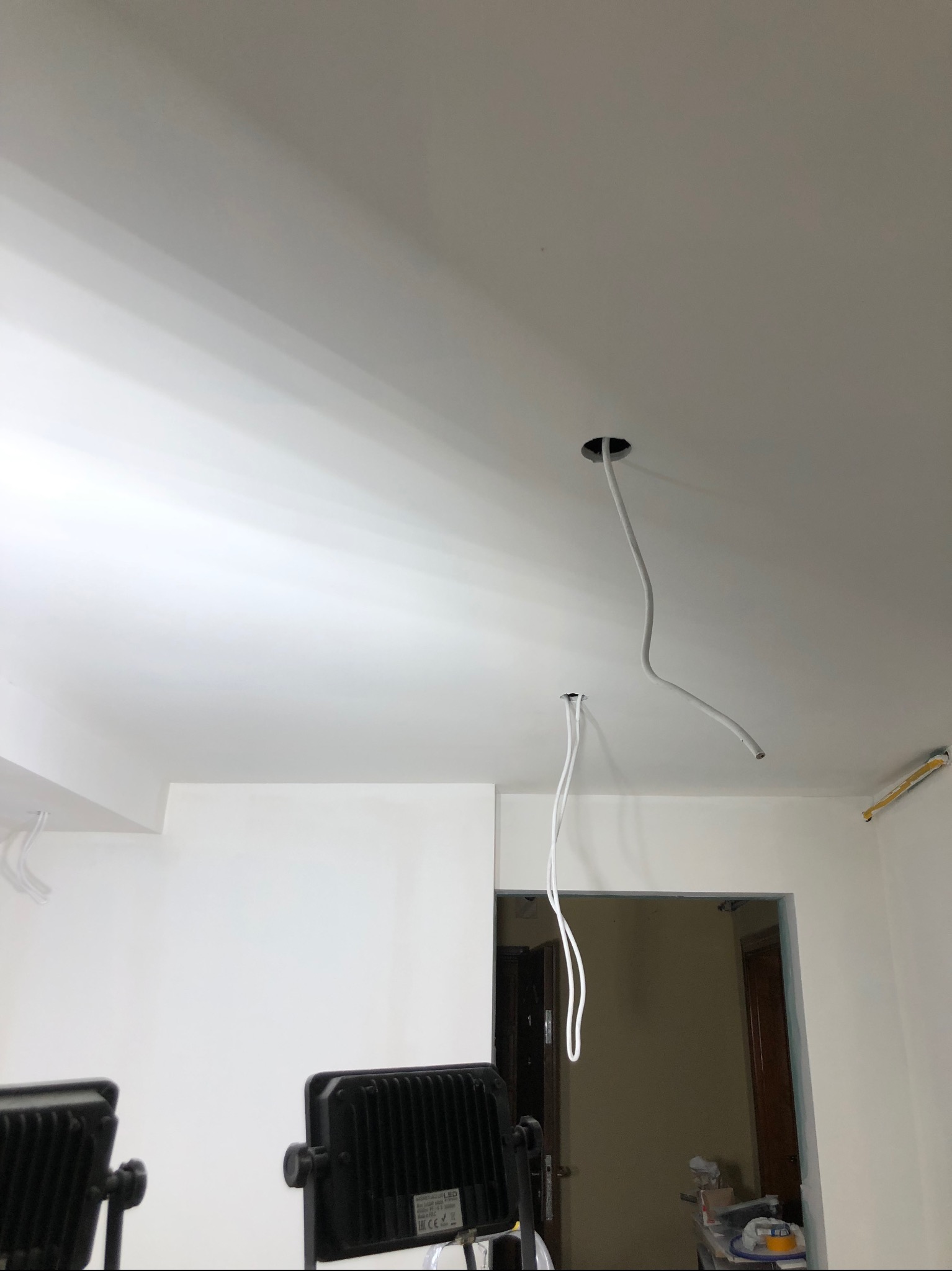 Biały sufit z dwoma otworami, z których zwisają kable elektryczne; widoczne również lampy budowlane i fragment ościeżnicy drzwiowej.