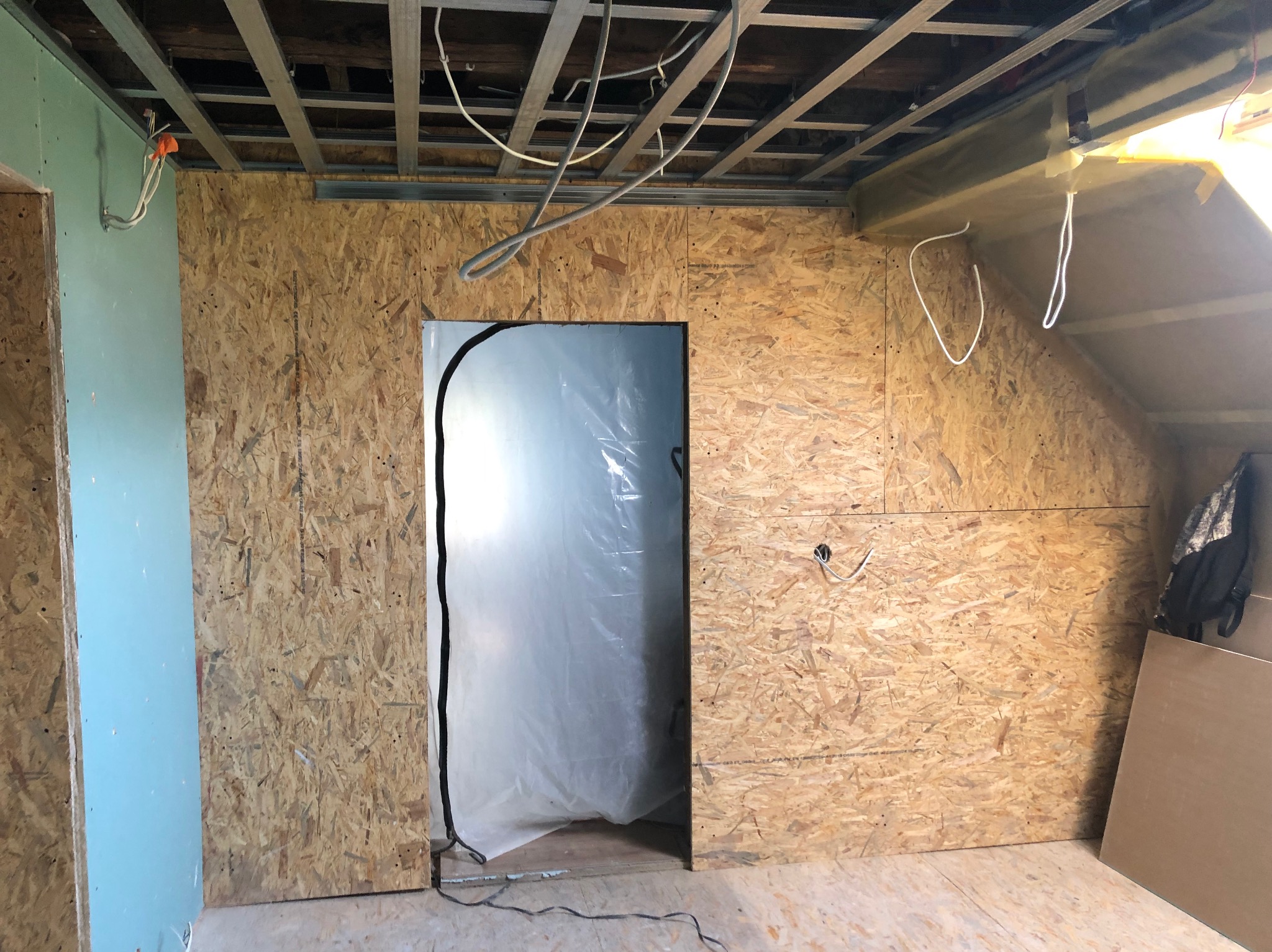 Wnętrze poddasza w trakcie wykańczania: ściany z płyt OSB, widoczne kable elektryczne, metalowa konstrukcja sufitu podwieszanego, drzwi zabezpieczone folią malarską, fragment ściany z płyty...