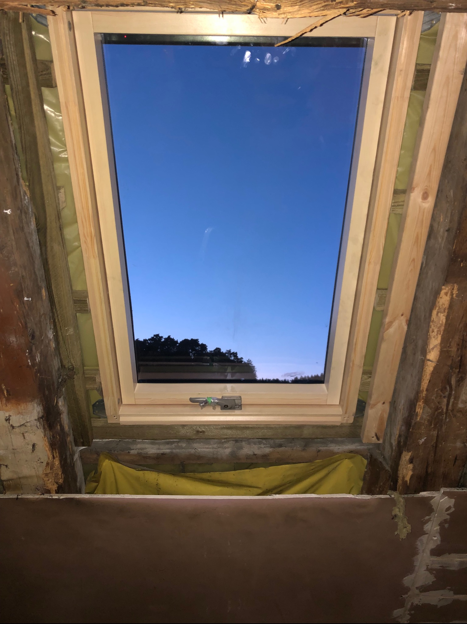 Widok od wewnątrz na nowe okno dachowe z drewnianą ramą, z widocznym niebieskim niebem o zmierzchu, w trakcie montażu na poddaszu z surowym drewnem i izolacją.