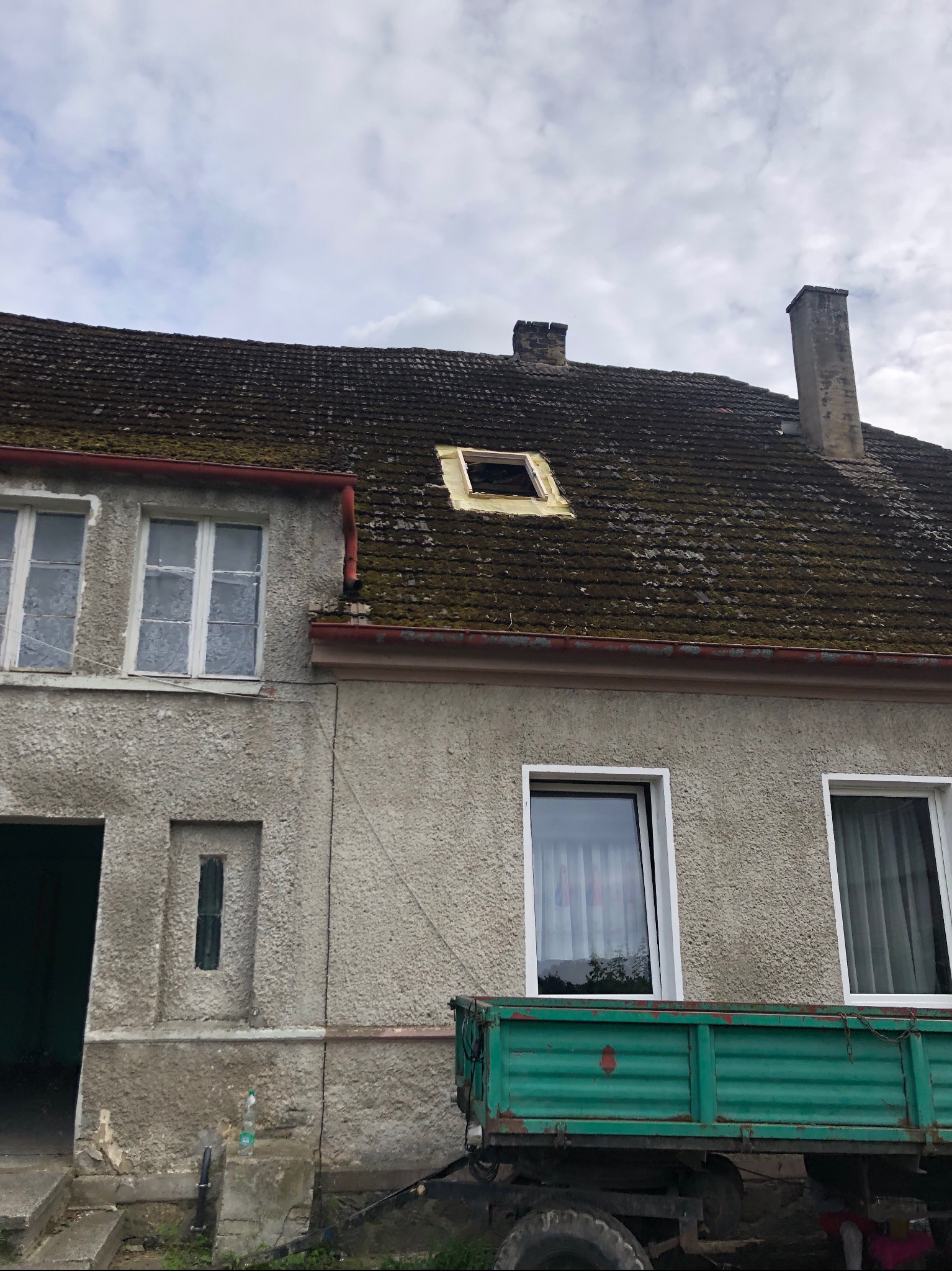 Stary, zaniedbany dom z dachem pokrytym mchem i brakującymi dachówkami, widoczny fragment zielonej przyczepy samochodu ciężarowego.