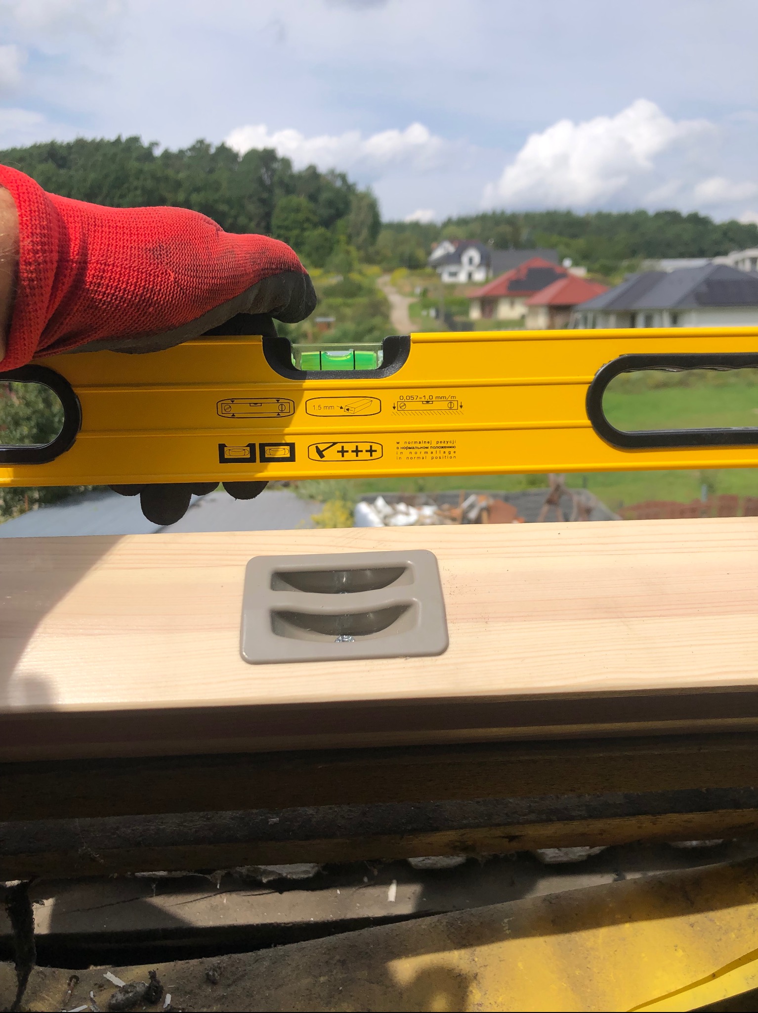 Poziomica w dłoni w czerwonej rękawicy, sprawdzająca wypoziomowanie jasnej deski z szarym uchwytem na tle zieleni i zabudowań.