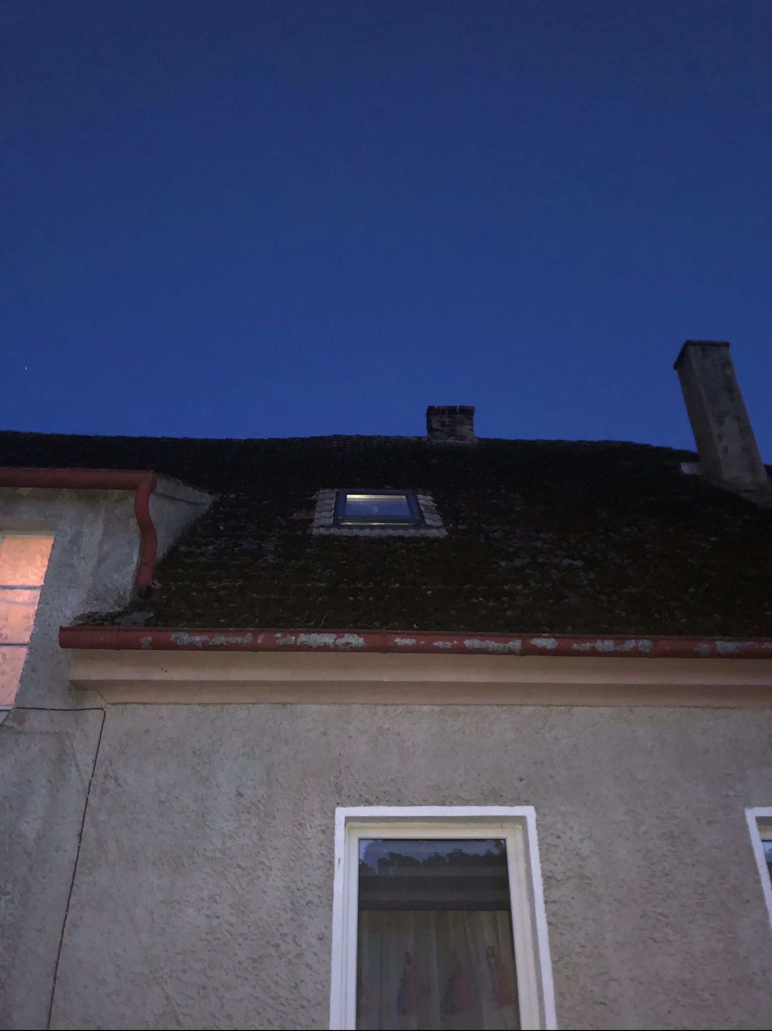 Niski kąt widzenia na dach pokryty mchem z oknem dachowym i kominem, widoczne orynnowanie i fragment elewacji budynku w wieczornym świetle.