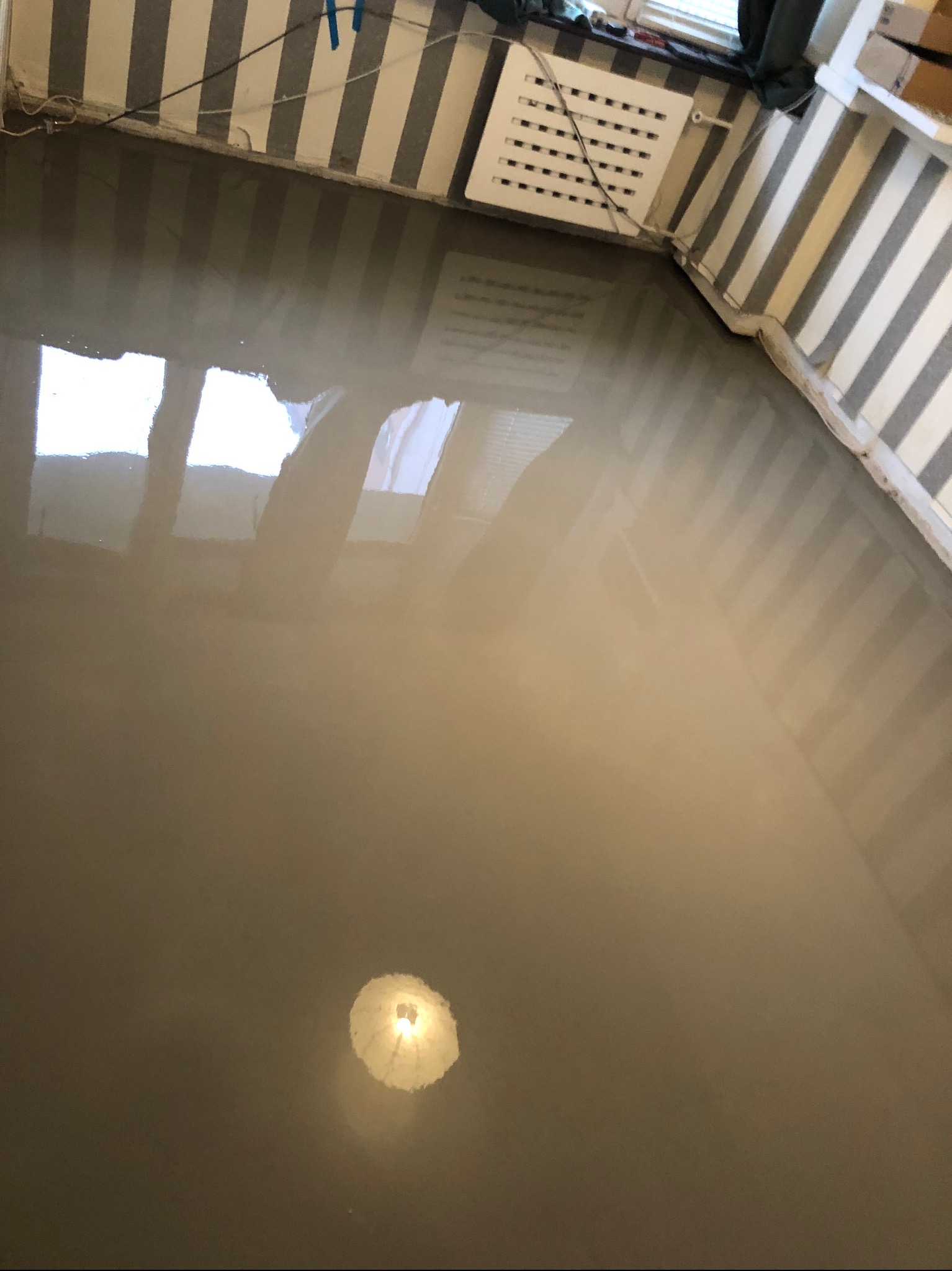 Świeżo wylana, samopoziomująca wylewka na podłodze w pomieszczeniu z paskowanymi ścianami, widoczne odbicie okna i lampy w mokrej powierzchni.