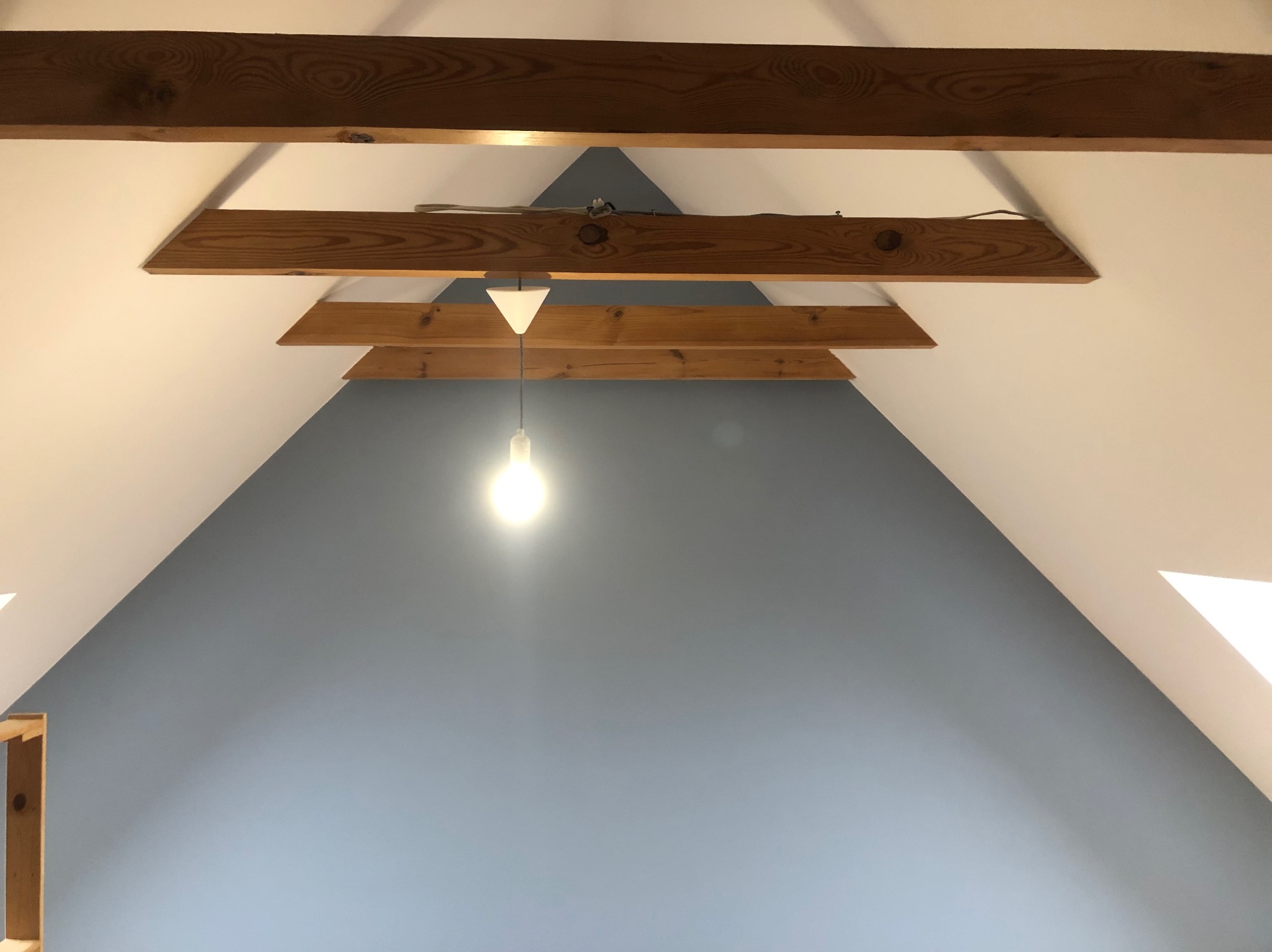 Poddasze z drewnianymi belkami stropowymi, pomalowane na biało i szaro-niebiesko, z wiszącą żarówką z białym kloszem.