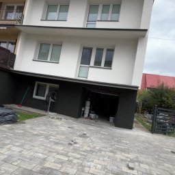 Usługi ogólnobudowlane - Dwukondygnacyjny dom z białymi ścianami i ciemną dolną częścią elewacji w trakcie prac wykończeniowych przy oknie. Na podjeździe kostka brukowa.