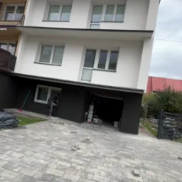 Dwukondygnacyjny dom z białymi ścianami i ciemną dolną częścią elewacji w trakcie prac wykończeniowych przy oknie. Na podjeździe kostka brukowa.