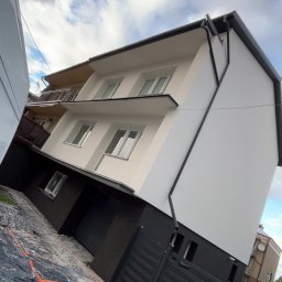 Usługi ogólnobudowlane - Ocieplony budynek z wykończoną elewacją w odcieniach bieli i szarości z czarnymi rynnami. Na pierwszym planie zabezpieczenia na chodniku.