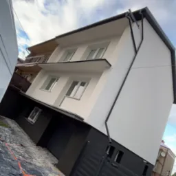 Ocieplony budynek z wykończoną elewacją w odcieniach bieli i szarości z czarnymi rynnami. Na pierwszym planie zabezpieczenia na chodniku.