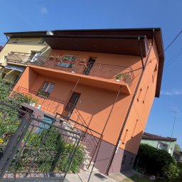 Usługi ogólnobudowlane - Pomarańczowy dom z balkonami i brązowym dachem w słoneczny dzień, widziany z poziomu gruntu, z otwartą furtką i elementami zieleni w kadrze.