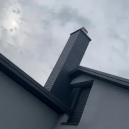 Szary dom z ciemnym dachem i kominem w obiektywie. Kąt ujęcia podkreśla geometrię budynku na tle pochmurnego nieba.