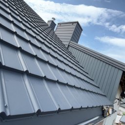 Usługi ogólnobudowlane - Grafitowy dach z blachodachówki modułowej, kominy w kolorze dachu na tle błękitnego nieba. Realizacja w Zawistach Podleśnych.