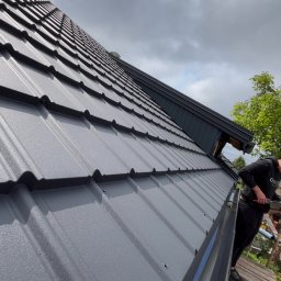 Usługi ogólnobudowlane - Szary, metalowy dach z bliska, z widocznym fragmentem orynnowania i pracownikiem na rusztowaniu, na tle pochmurnego nieba i zieleni drzew.