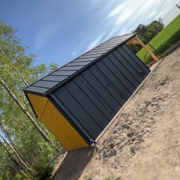 Usługi ogólnobudowlane - Gotowy, nowoczesny domek narzędziowy z czarną blachą i żółtymi detalami, usytuowany na trawiastym terenie pod błękitnym niebem. Widoczne drzewa i pola w tle.