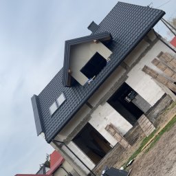 Usługi ogólnobudowlane - Dom w stanie surowym z czarną dachówką, widoczne elementy konstrukcyjne i okna dachowe. Ujęcie z dołu, podkreślające geometrię dachu i bryłę budynku.