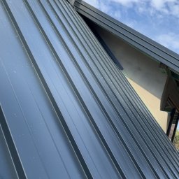 Usługi ogólnobudowlane - Szary dach z blachy trapezowej, widok pod kątem. Na dachu komin wentylacyjny w kolorze dachu. W tle błękitne niebo z chmurami.