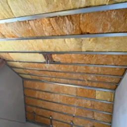 Ocieplenie poddasza wełną mineralną, widok od dołu. Konstrukcja metalowa z wypełnieniem z izolacji, fragment ściany. Widoczny etap prac wykończeniowych.