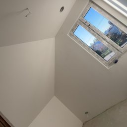 Usługi ogólnobudowlane - Poddasze po tynkowaniu na biało, widoczne okno dachowe z widokiem na zewnątrz i wystające kable. Ściany gładkie, przygotowane do dalszych prac wykończeniowych.