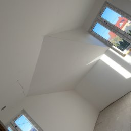 Usługi ogólnobudowlane - Białe poddasze po remoncie: skośny sufit z oknami dachowymi, ściany przygotowane do malowania. Widoczne gniazdka elektryczne i fragment drewnianej konstrukcji.