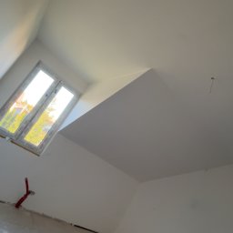 Usługi ogólnobudowlane - Poddasze po remoncie: gładkie, białe ściany, okno dachowe wpuszczające światło, widoczne instalacje. Świeżo wykończone wnętrze z ukośnym sufitem.