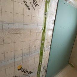 Usługi ogólnobudowlane - Montaż folii paroizolacyjnej Isover Vario XtraSafe z taśmą Vario MultiTape+ przy płycie karton-gips. Widoczne dłonie osoby dociskającej taśmę. Ocieplenie wełną mineralną w tle.