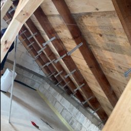 Usługi ogólnobudowlane - Poddasze w trakcie prac: drewniana konstrukcja dachu z zamocowanymi metalowymi elementami, widoczne przewody i narzędzia. Przygotowanie do ocieplenia.