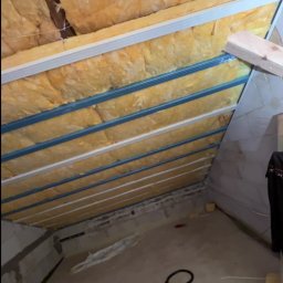 Usługi ogólnobudowlane - Skośne poddasze z ociepleniem z wełny mineralnej, widoczne metalowe profile konstrukcyjne i fragment betonowej ściany. Ujęcie z dołu, widać podłogę i elementy instalacji.