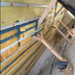 Usługi ogólnobudowlane - Skośny kadr na ocieplone wełną mineralną poddasze z widoczną konstrukcją drewnianą i metalowymi profilami. Fragment kartonu z rurami na podłodze.