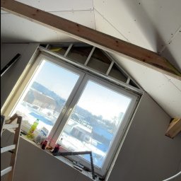 Usługi ogólnobudowlane - Poddasze z oknem dachowym w trakcie wykańczania płytami. Widoczne drewniane belki konstrukcyjne i narzędzia pracy. Prace wykończeniowe w jasnym wnętrzu.