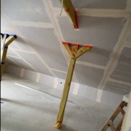 Usługi ogólnobudowlane - Poddasze w trakcie wykańczania: widoczne płyty gipsowe na skosach, drewniane podpory zabezpieczone folią, drabina. Prace wykończeniowe w surowym wnętrzu.