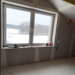 Usługi ogólnobudowlane - Poddasze z oknem po montażu płyt gipsowo-kartonowych, widoczne narzędzia na parapecie i taśma zabezpieczająca. Za oknem zimowy krajobraz.