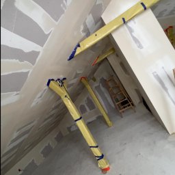 Usługi ogólnobudowlane - Skośny sufit i ściany po szpachlowaniu, zabezpieczone rury, drabina w tle. Prace wykończeniowe w pomieszczeniu o nieregularnym kształcie.